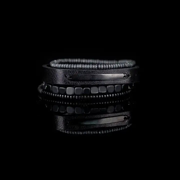 Black multi-layer men’s bracelet set featuring matte onyx beads, braided black rope, crystal cross centerpiece, gold chevron metal bar, and layered metallic accents on a light background.