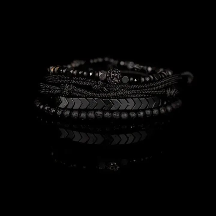 Black layered men’s bracelet set with chevron metal strand, lava stone beads, faceted metallic accents, braided rope wraps, and pavé centerpiece on white background.