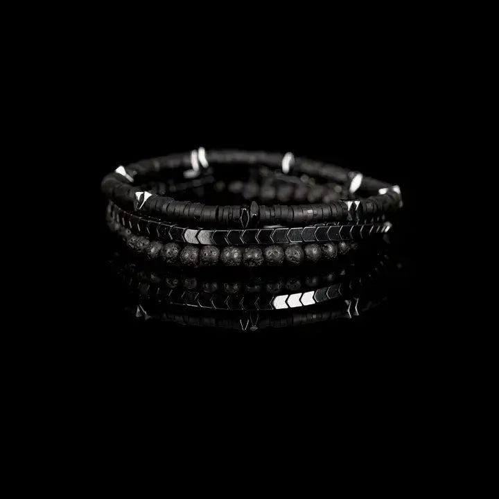 Black multi-layer men’s bracelet featuring hematite disc strands, shield-pattern metallic beads, pyramid-cut spacers, and lava stone bead accents on a light background.