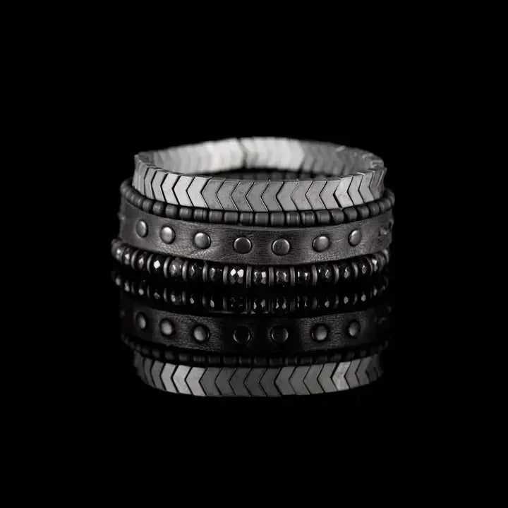 Men’s layered bracelet set featuring a silver-tone chevron stretch bracelet, black leather cuff with gunmetal stud details, matte black bead strand, and faceted black stone beads, stacked in a rugged contemporary luxury design on a white background.