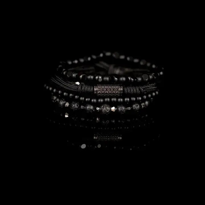 Black layered men’s bracelet set with matte black beads, lava stone accents, faceted geometric beads, braided rope wraps, and pavé cylindrical centerpiece on white background.