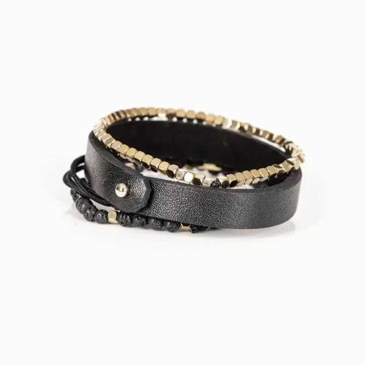 Men’s layered bracelet set featuring a black textured leather cuff, gold-tone faceted metal bead bracelet, black braided cord wrap, matte black lava stone beads, and gold-tone accent beads, stacked in a refined modern luxury design on a white background.