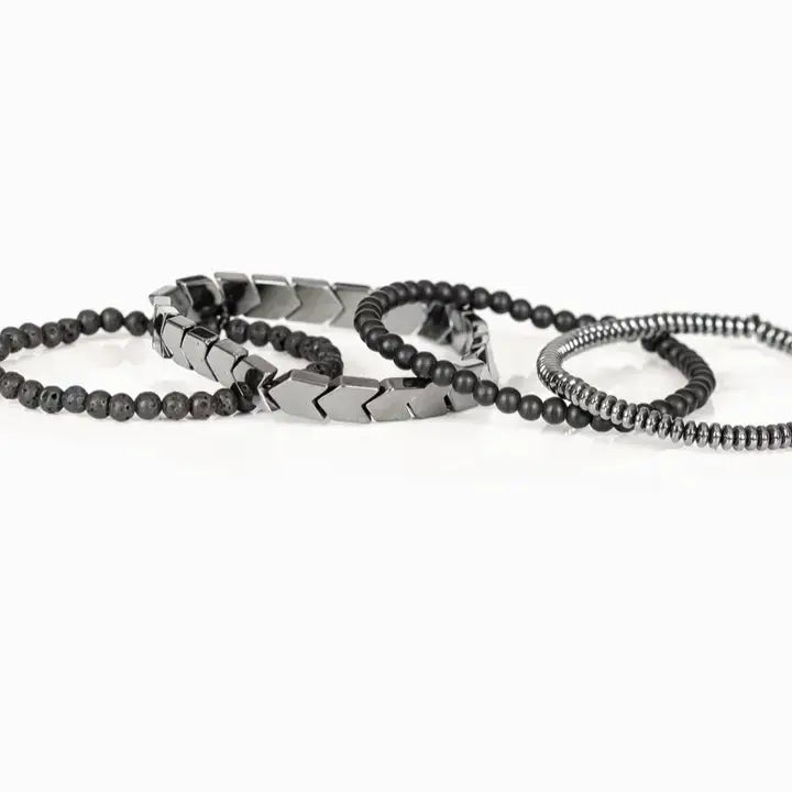 Men’s layered bracelet set featuring gunmetal chevron metal beads, matte black lava stone beads, black matte bead strands, and silver-tone hematite spacer beads, stacked in a bold industrial luxury design on a white background.
