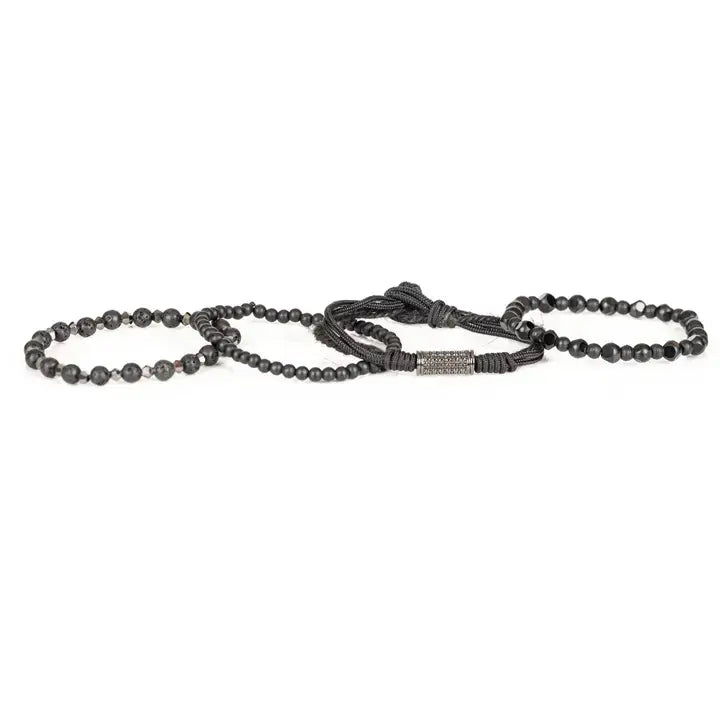 Black layered men’s bracelet set with matte black beads, lava stone accents, faceted geometric beads, braided rope wraps, and pavé cylindrical centerpiece on white background.