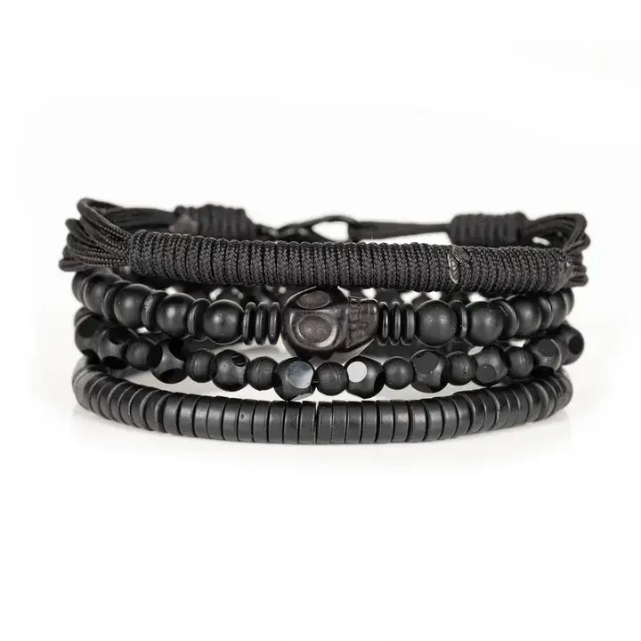 Men's black layered bracelet set featuring braided cord, matte black beads, faceted black crystal beads, a carved skull centerpiece, and black heishi disc beads, arranged in a stacked design on a white background.