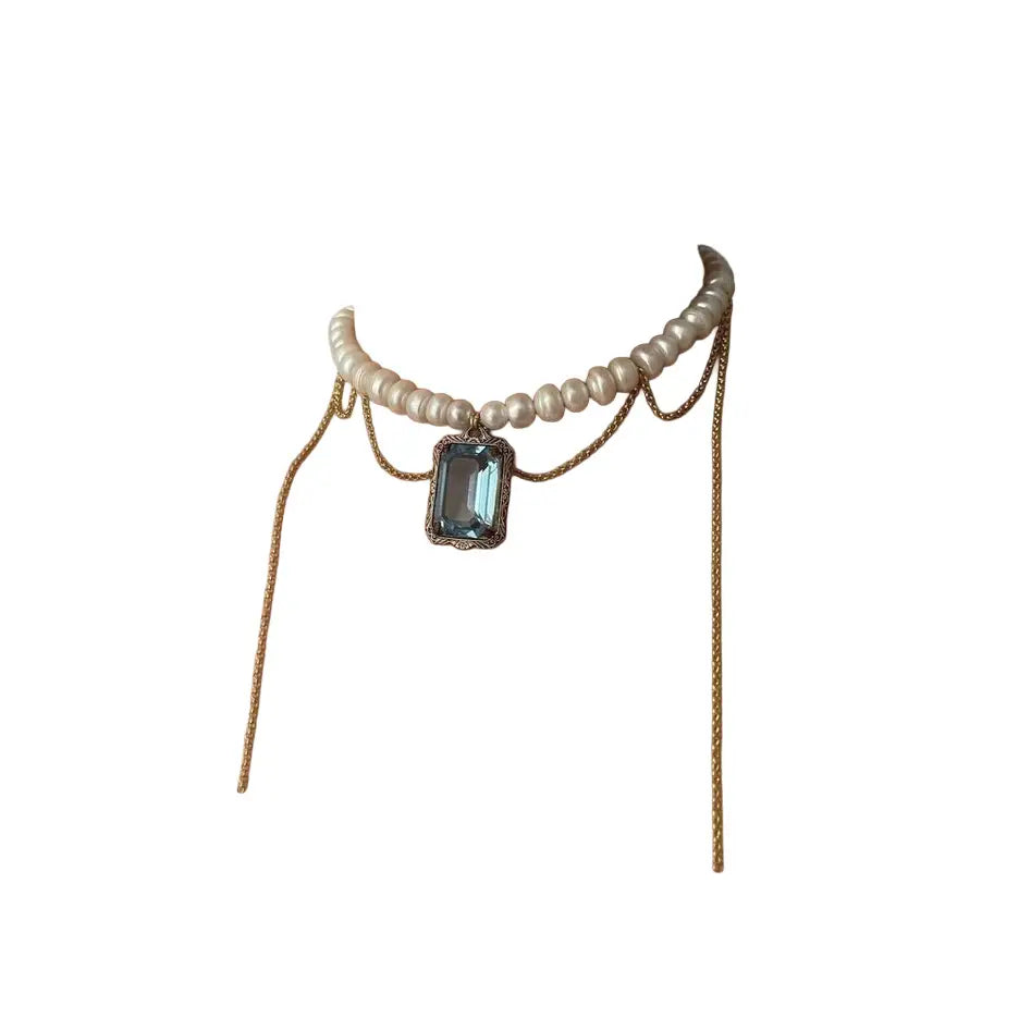 Pearl necklace with draped gold chains and a blue crystal pendant on a white background.