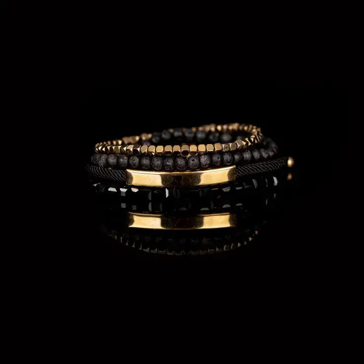 Men’s layered bracelet set featuring faceted black crystal beads, matte black lava stone beads, black braided cord, a polished gold-tone bar centerpiece, and gold-tone hematite-style spacer beads, stacked in a sophisticated black and gold luxury design on a white background.