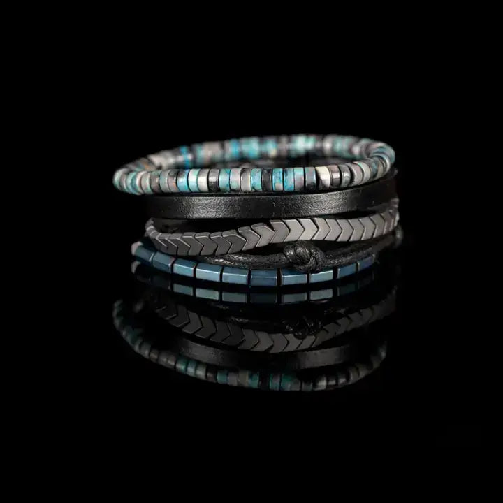 Blue and black multi-layer men’s bracelet set featuring silver chevron metal bar, blue-grey stone beads, black leather band, braided cord, and metallic hematite cube beads on a light background.
