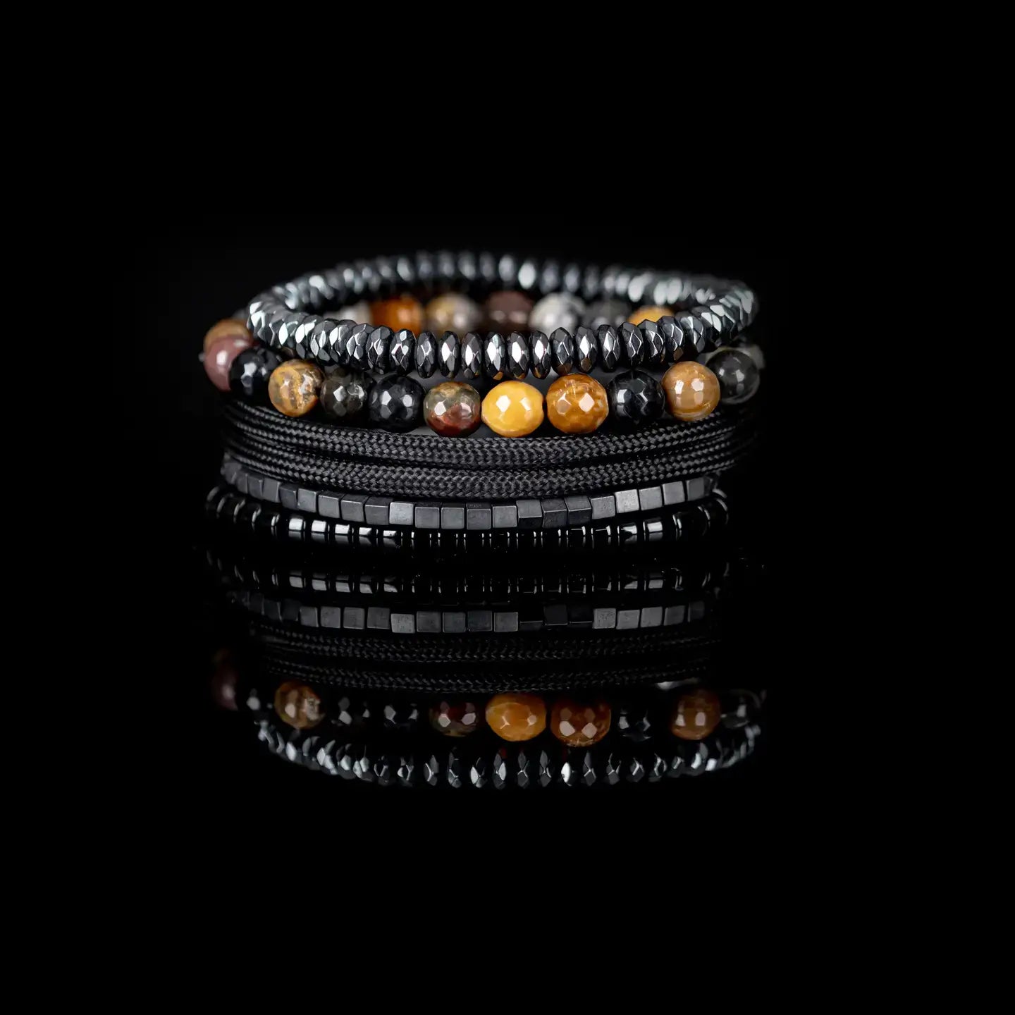 Black multi-layer men’s bracelet featuring natural multi-tone stone beads, faceted hematite strands, metallic cube accents, and braided black rope layers on a light background.