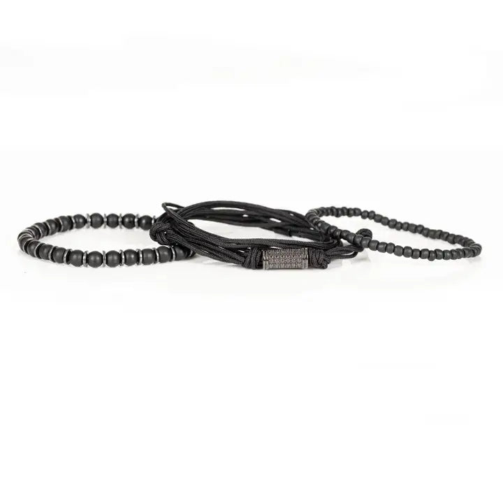 Black layered men’s bracelet set with matte black beads, braided rope wraps, metallic spacer accents, and pavé cylindrical centerpiece on white background.