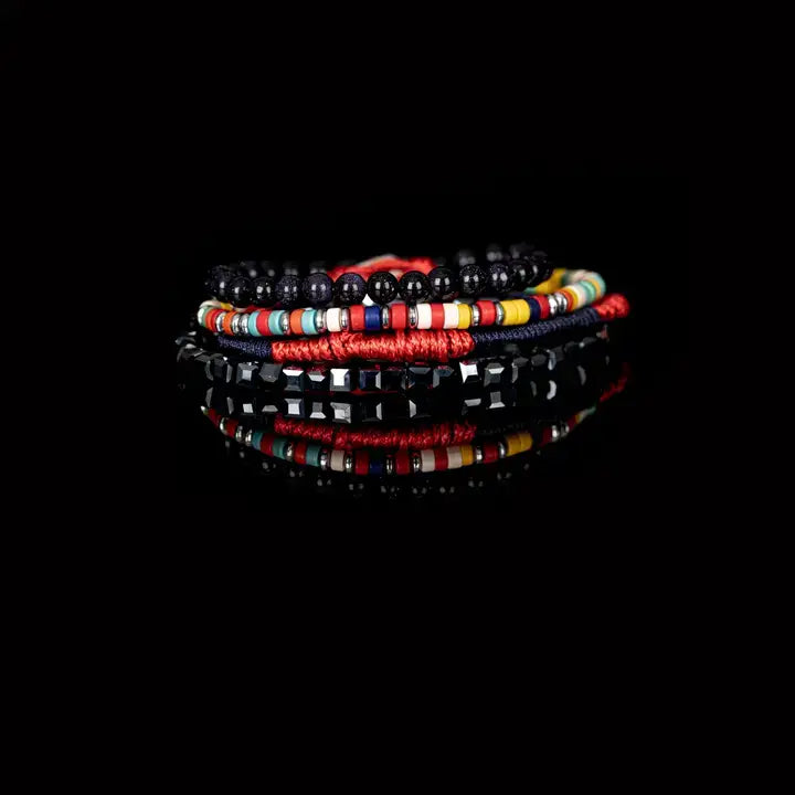 Men’s layered bracelet set featuring glossy black onyx-style beads, multicolor seed beads, red woven cord, navy braided cord, and faceted hematite cube beads, stacked in a bold contemporary luxury design on a white background.