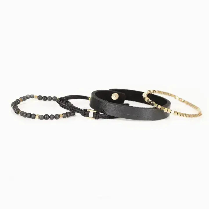 Men’s layered bracelet set featuring a black textured leather cuff, gold-tone faceted metal bead bracelet, black braided cord wrap, matte black lava stone beads, and gold-tone accent beads, stacked in a refined modern luxury design on a white background.