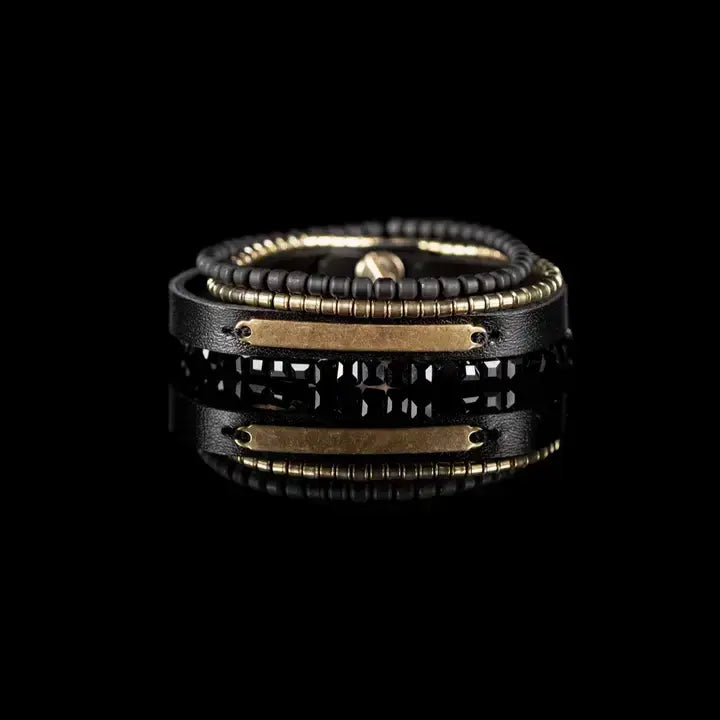 Men’s layered bracelet set featuring a black leather cuff with a gold-tone ID plate, matte black bead strand, gold-tone heishi beads, and faceted black crystal beads, stacked in a sleek modern luxury design on a white background.